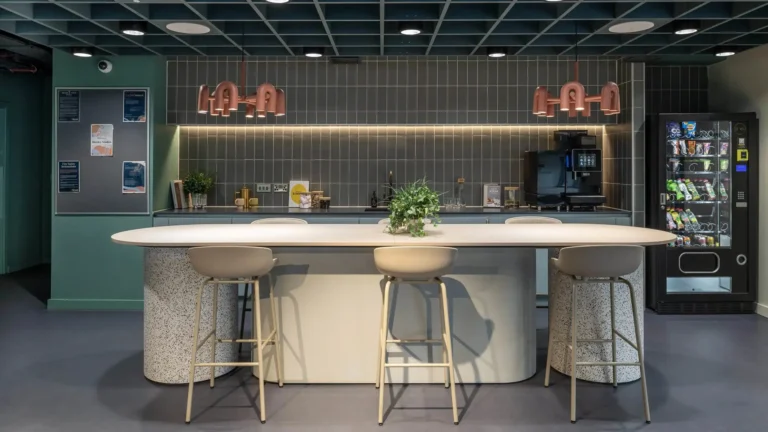 A kitchen and dining area. There is a breakfast bar with six stools around it, and behind that a kitchen unit with a coffee machine. To the right is a vending machine; to the left, a noticeboard