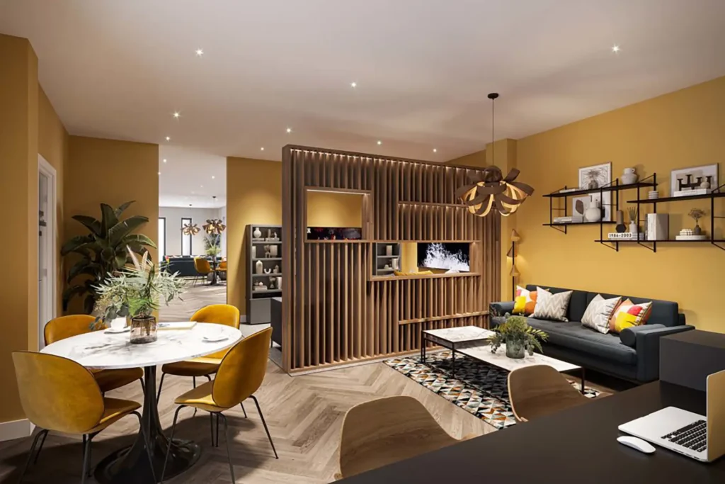 A large room in student accommodation. In the foreground there is a desk; in the middle distance a dining table with chairs, and a sofa; in the background, more seating and some shelving