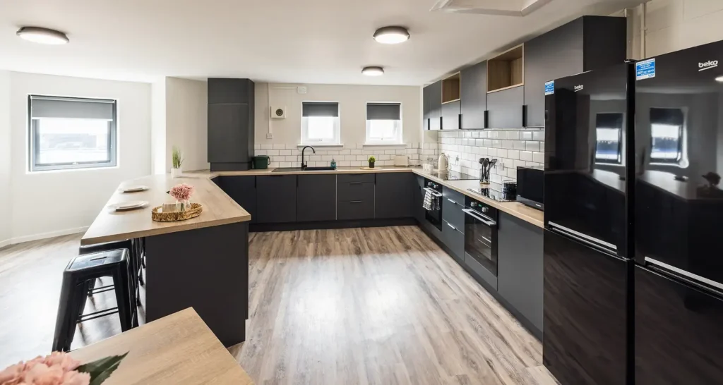 A large shared kitchen. It is well equipped with two fridge freezers, two ovens, a sink, and a microwave. There are work surfaces and a breakfast bar with stools