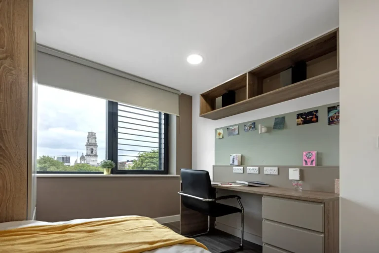 A bedroom at Stanhope House. There is a large window, creating a light filled room, with a view of Portsmouth. In the room there is a large desk with storage space and, at the front of the picture, a bed.