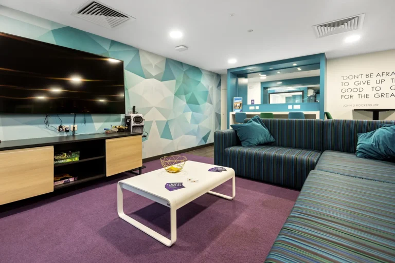 A shared living space with a large sofa, a coffee table and a large wall-mounted TV In the background is a hight workbench with three chairs.