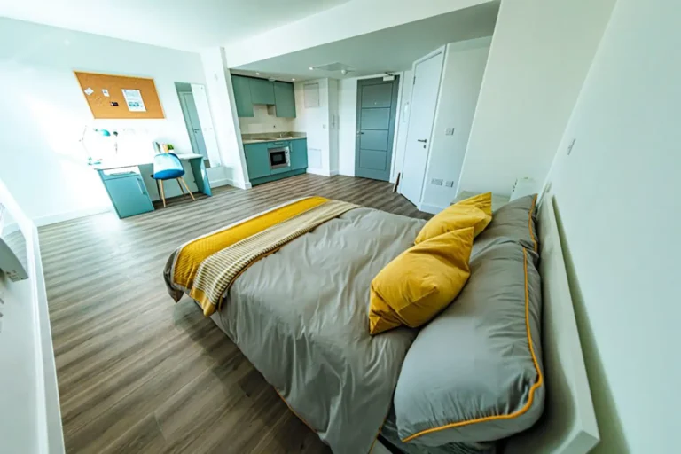 A large student bedroom/ There is a large bed in the foreground, with a desk and kitchen area in the background