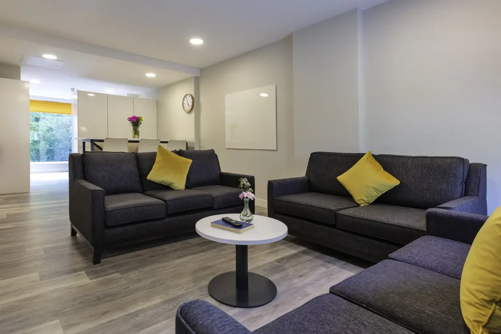 A communal living space. Three seater sofas are positioned around a coffee table. In the background is a breakfast bar area with high stools. The room is light.