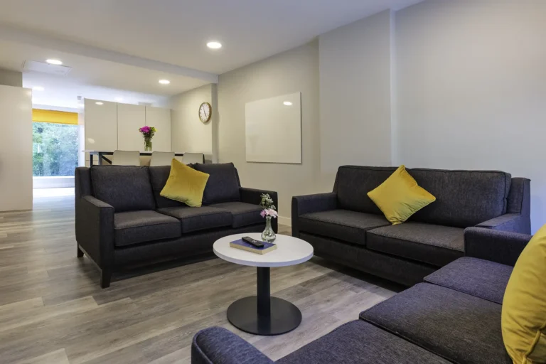A communal living space. Three seater sofas are positioned around a coffee table. In the background is a breakfast bar area with high stools. The room is light.
