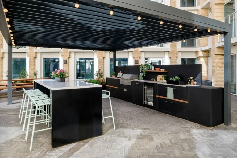 A large outdoor eating area, with a high table with chairs around it. and a large outdoor kitchen facility including a grill, fridge and food prep area. The area is covered by a canopy.