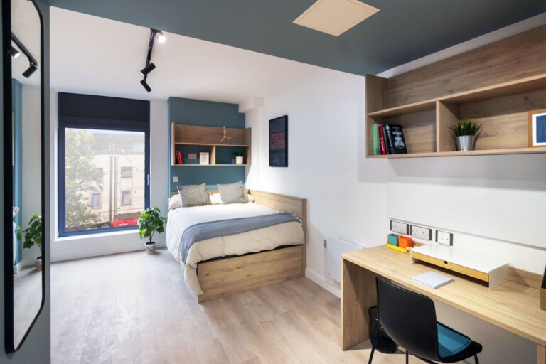 A student bedroom. It has shelving units, a bed, and a desk. The room is light and spacious