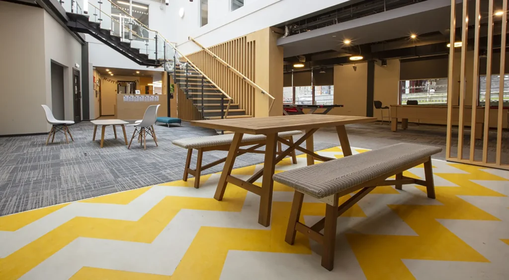 A communal area at Fraser Studios. In the foreground is a large table with benches, behind that a smaller table with a few chairs around it and a staircase leading up.