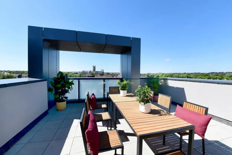 A rooftop social space. A large table with seven seats around it. In the background is Canterbury Cathedral against a glorious blue sky.