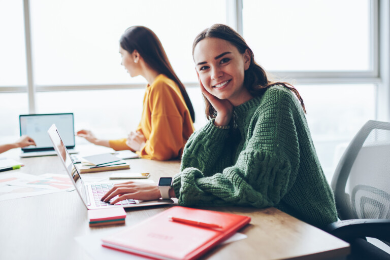 Happy female university student looking at the camera whilst she is studying , using a laptop and books