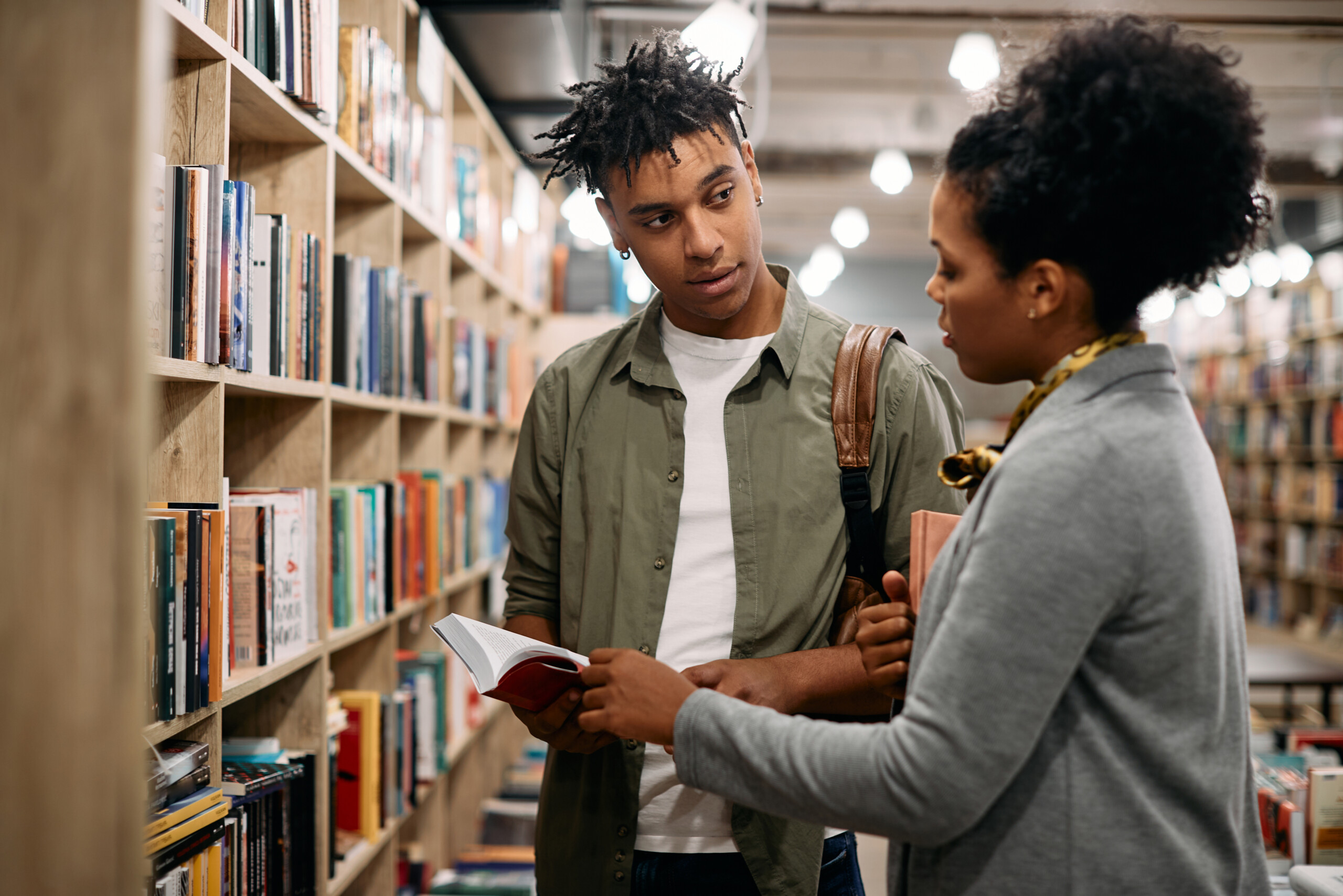 Young student speaking to a librarian in a library, talking about a book the librarian is holding