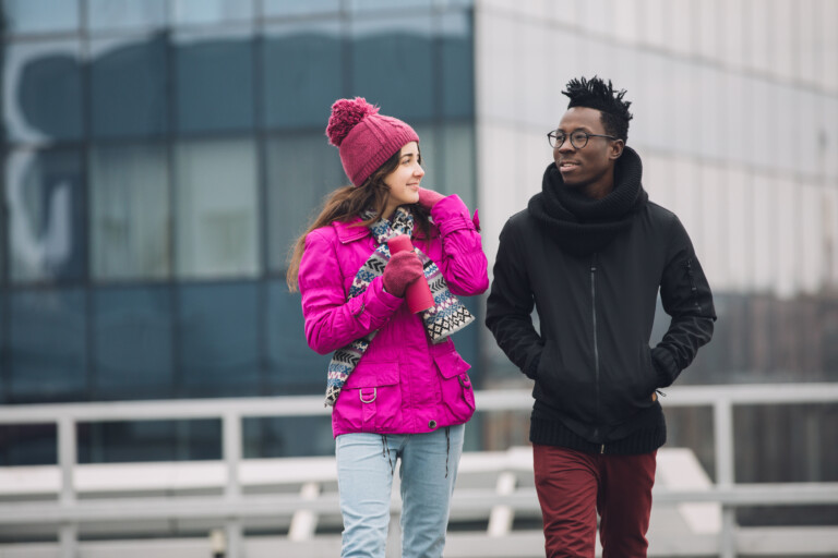 Two friends on a walk bundled up in coats and hats. They are laughing and walking. The girl is wearing a bright pink coat with matching flask. The guy is wearing a black coat