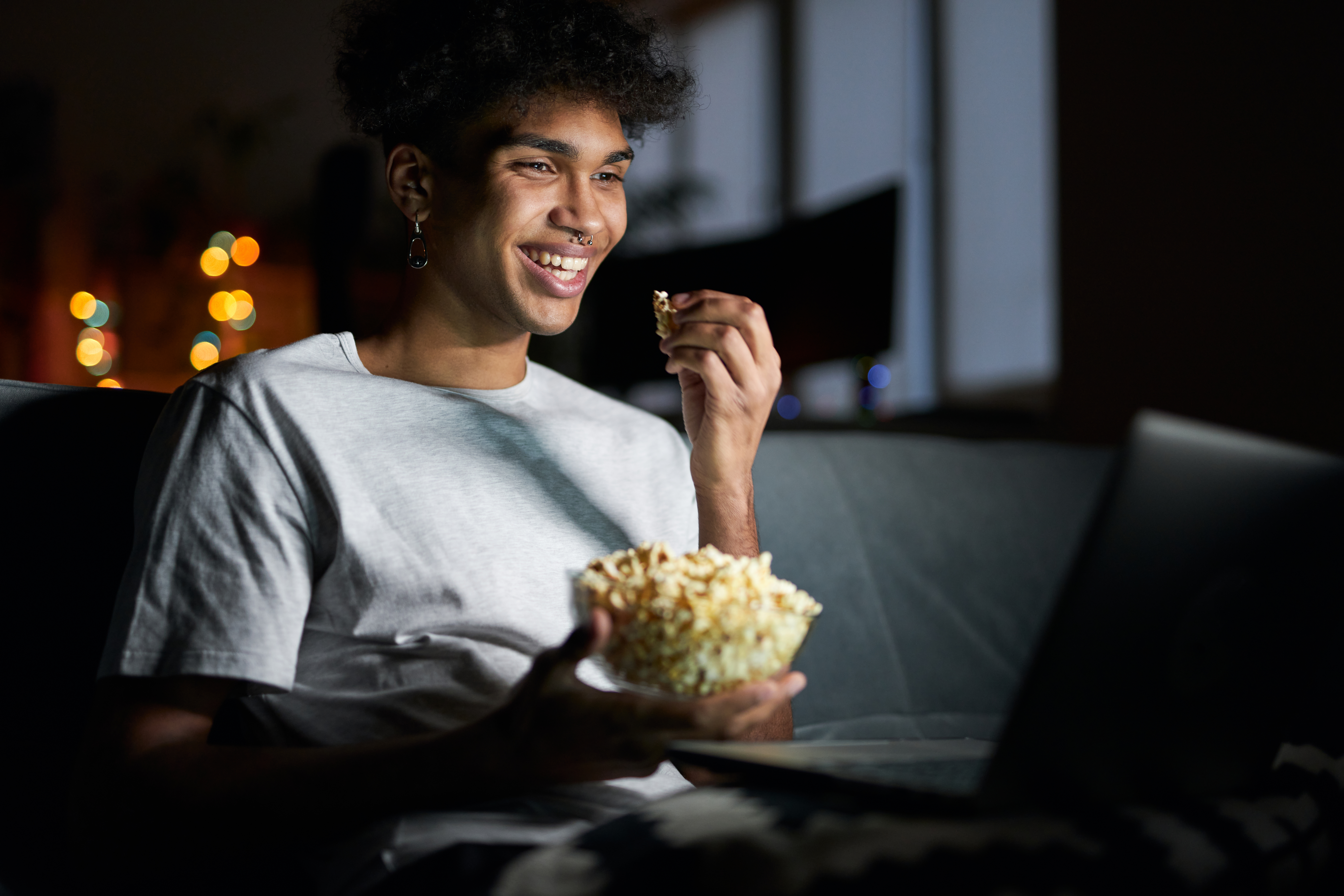 Happy young guy or student sitting on his sofa at home. He is eating popcorn and laughing at a movie he is watching on his laptop.